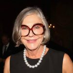 زنی مسن با موهای خاکستری کوتاه، عینک بزرگ قهوهای و گردنبند مروارید که در یک فضای رسمی و نور کم لبخند میزند.