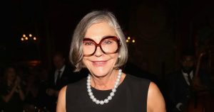 زنی مسن با موهای خاکستری کوتاه، عینک بزرگ قهوه‌ای و گردنبند مروارید که در یک فضای رسمی و نور کم لبخند می‌زند.