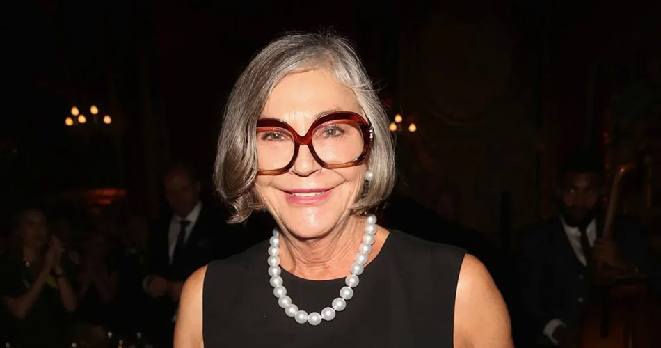 زنی مسن با موهای خاکستری کوتاه، عینک بزرگ قهوهای و گردنبند مروارید که در یک فضای رسمی و نور کم لبخند میزند.