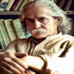 مردی میانسال با موهای بلند خاکستری و سبیل پرپشت نشسته در کتابخانه، در حالی‌که میان قفسه‌های کتاب احاطه شده است.