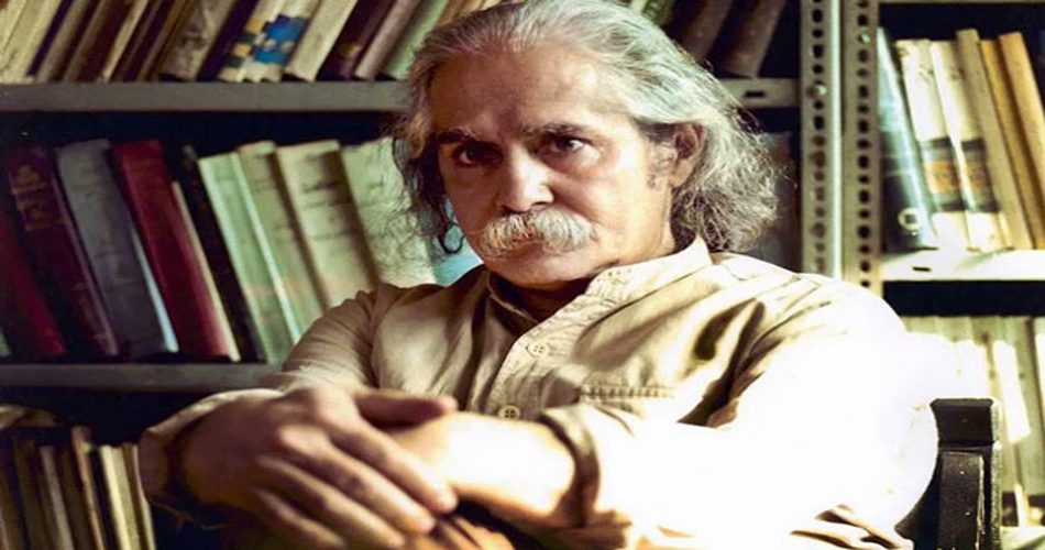 مردی میانسال با موهای بلند خاکستری و سبیل پرپشت نشسته در کتابخانه، در حالی‌که میان قفسه‌های کتاب احاطه شده است.