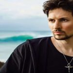 مردی جوان با ته‌ریش و گردنبند نقره‌ای در فضای ساحلی، در حال نگاه کردن به دوردست با پس‌زمینه دریا و صخره.