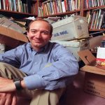 یک فرد در حال نشستن روی زمین در اتاقی پر از کتاب‌ها و جعبه‌های مقوایی، پشت سر او قفسه‌های پر از کتاب قرار دارد. فضای اتاق حالت قدیمی و ابتدایی دارد. نور نرم و طبیعی داخل اتاق پخش شده است.