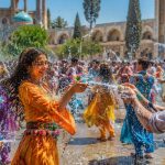 جمعی از جوانان با لباس‌های رنگارنگ در حیاطی با معماری سنتی ایرانی زیر نور آفتاب به هم آب می‌پاشند؛ دختری با لباس نارنجی در حال خندیدن و مردی روبه‌رویش آب می‌پاشد، قطرات آب در هوا پخش شده‌اند